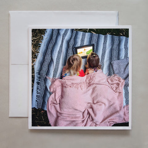 This greeting card shows two young children, cuddling and covered in blankets while watching an iPad, (being a typical Covid experience for our little ones!) by photographer Jennifer Echols.