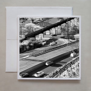 This black and white photo card shows cars on the Don Valley Parkway turnoff with the Evergreen Brickworks in the background.  Photographed from the Chester Hill Lookout by Toronto photographer, Caley Taylor.