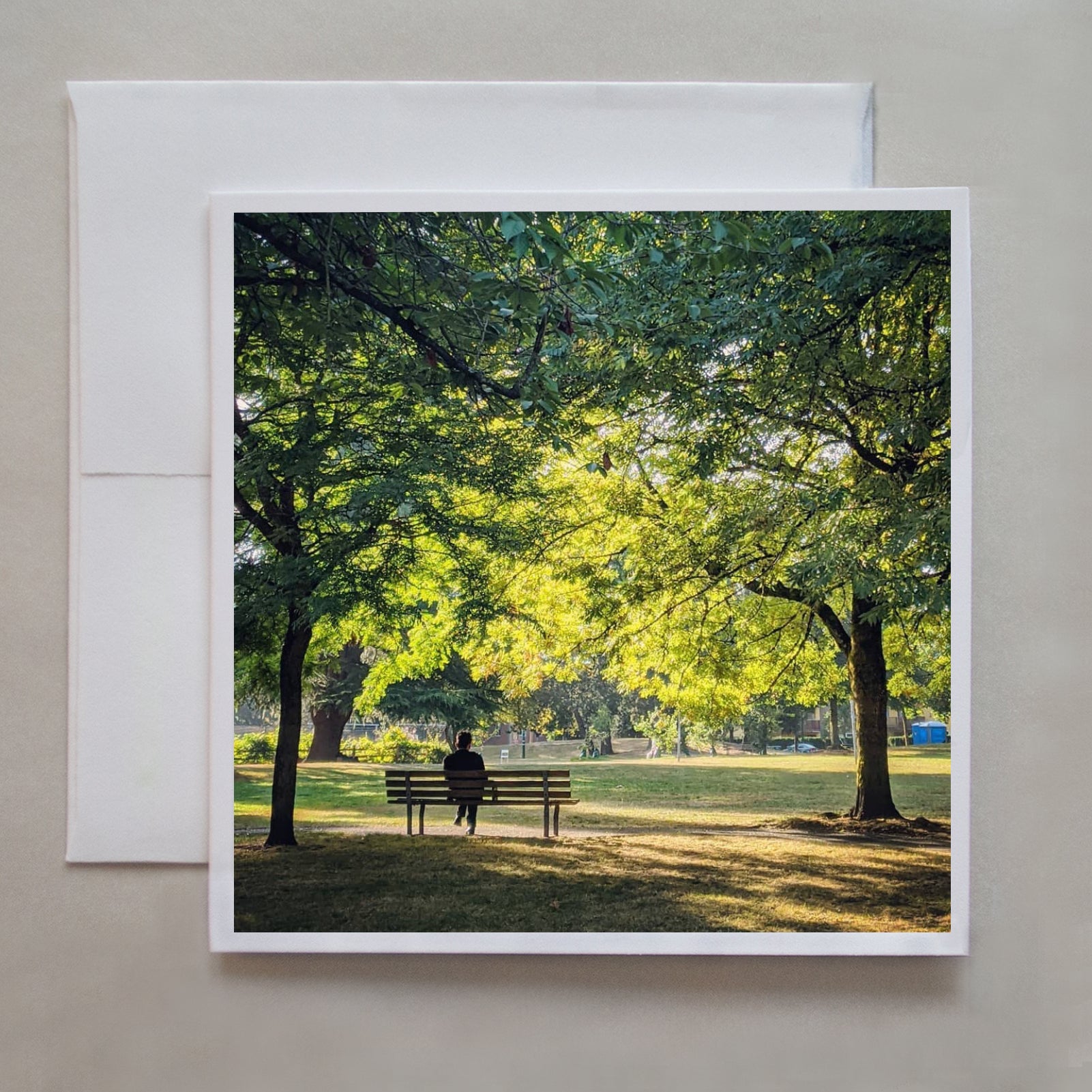 This notecard is of a person  reflecting on a park bench while sunlight drapes through summer-lush trees by photographer Jen Echols.