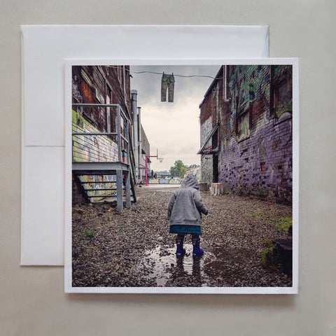 This greeting card is a darker, rainy-day photograph of a child puddling jumping amongst an urban landscape of walls covered in graffiti by photographer Jennifer Echols.