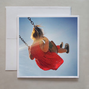 This summertime greeting card shows a little girl wearing a red dress, swinging in sunflares by photographer Jennifer Echols.