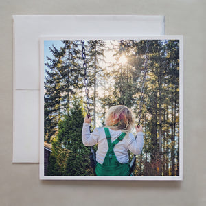 This greeting card shows a young child swinging amongst the trees and sunflares by photographer Jennifer Echols.